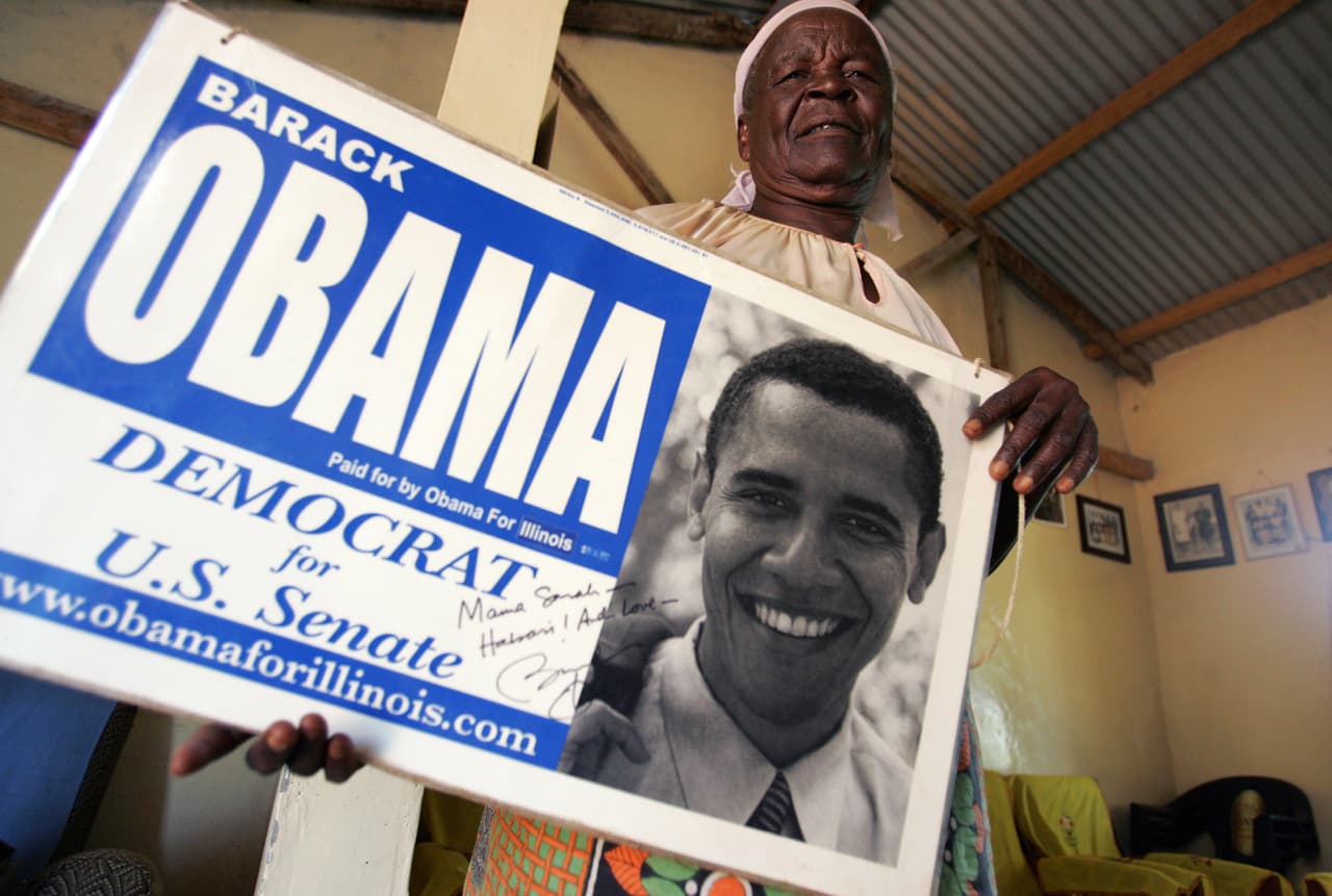 La abuela del expresidente con un cartel de campaña de Obama, en su casa de Kogelo, en febrero de 2008. “El fallecimiento de ‘Mamá Sarah’ es un gran golpe para nuestra nación. Hemos perdido a una mujer fuerte y virtuosa, una matriarca que mantuvo unida a la familia Obama y fue un ícono de los valores familiares”, dijo el presidente keniano Uhuru Kenyatta.
<br>