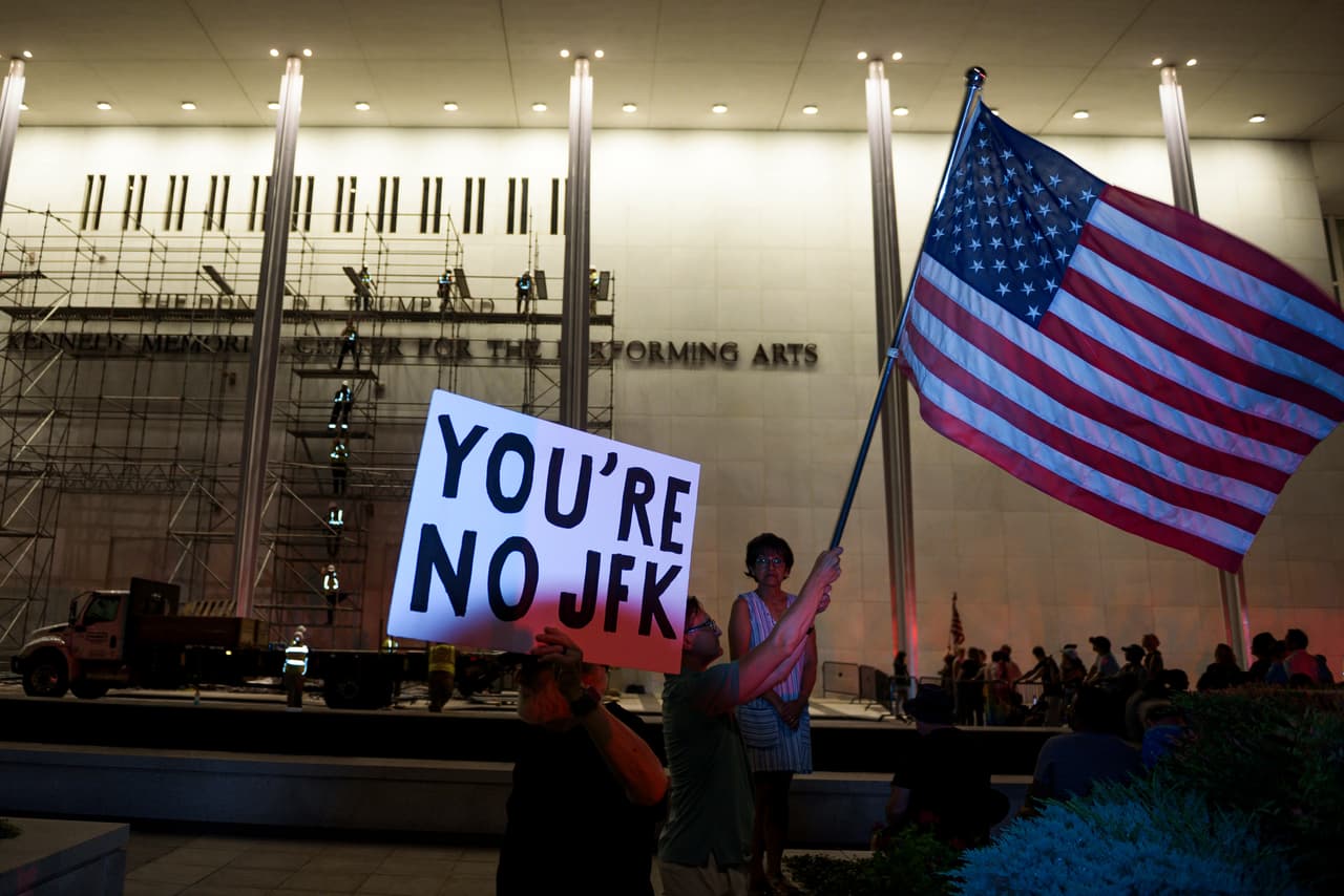 Manifestantes ondean la bandera estadounidense y pancartas mientras los trabajadores se preparan para retirar el nombre del presidente Donald Trump.