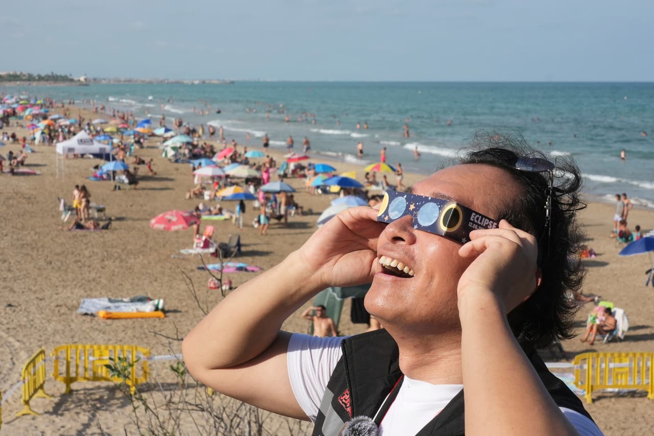 Un hombre lleva puestas unas gafas especiales para eclipses mientras los bañistas se reúnen antes del eclipse solar total, en una playa cerca de Valencia, España.