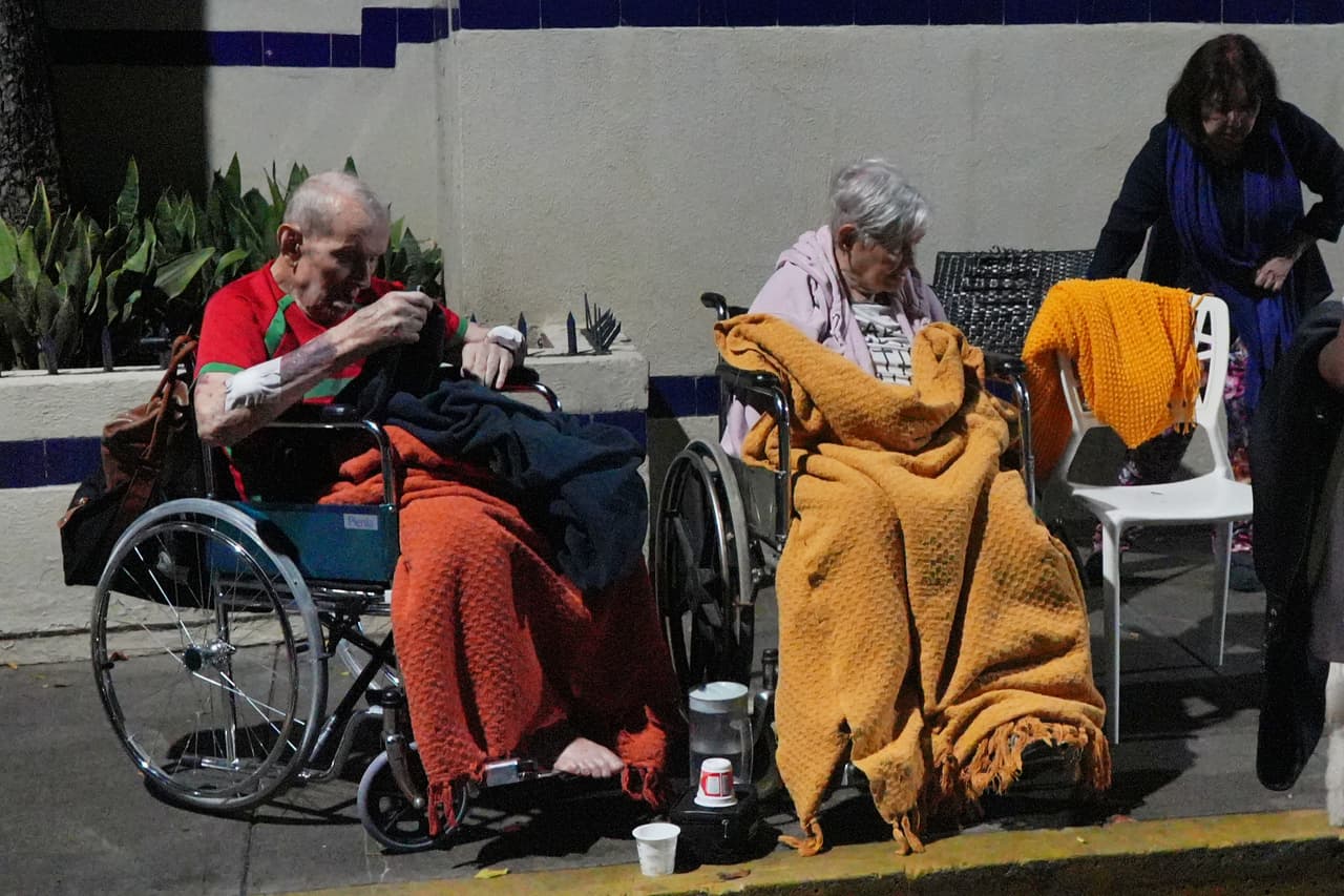 Personas mayores se sientan frente a edificios dañados tras un terremoto.