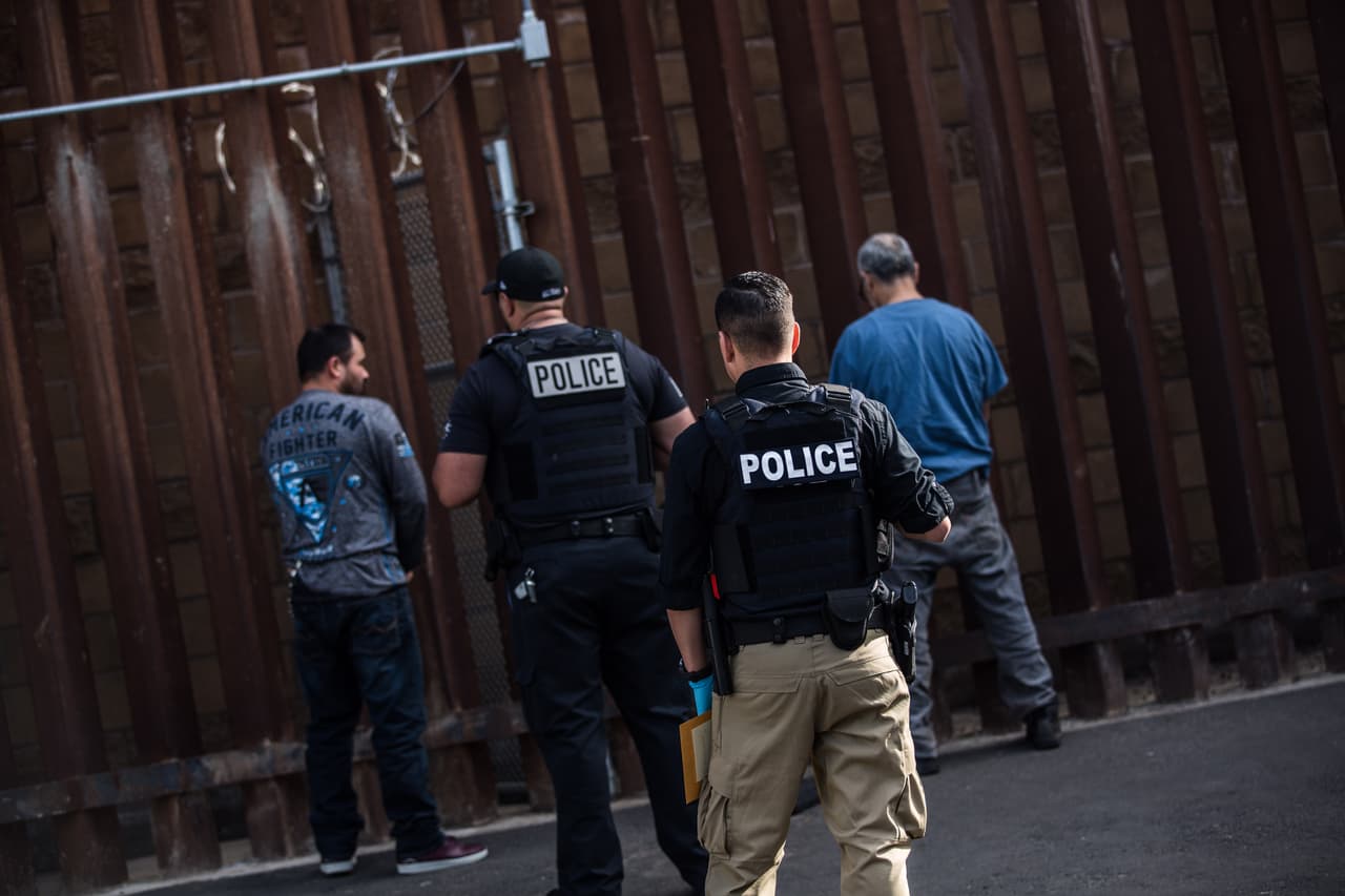 Dos policías de ICE vigilan a los dos detenidos mientras esperan que los oficiales mexicanos abran la puerta por donde se realizará la deportación. Ambos detenidos llevan cadenas que los amarran desde su cintura y terminan con unas esposas en sus manos. La espera dura unos quince minutos. Almudena Toral/Univision Digital
<br>
