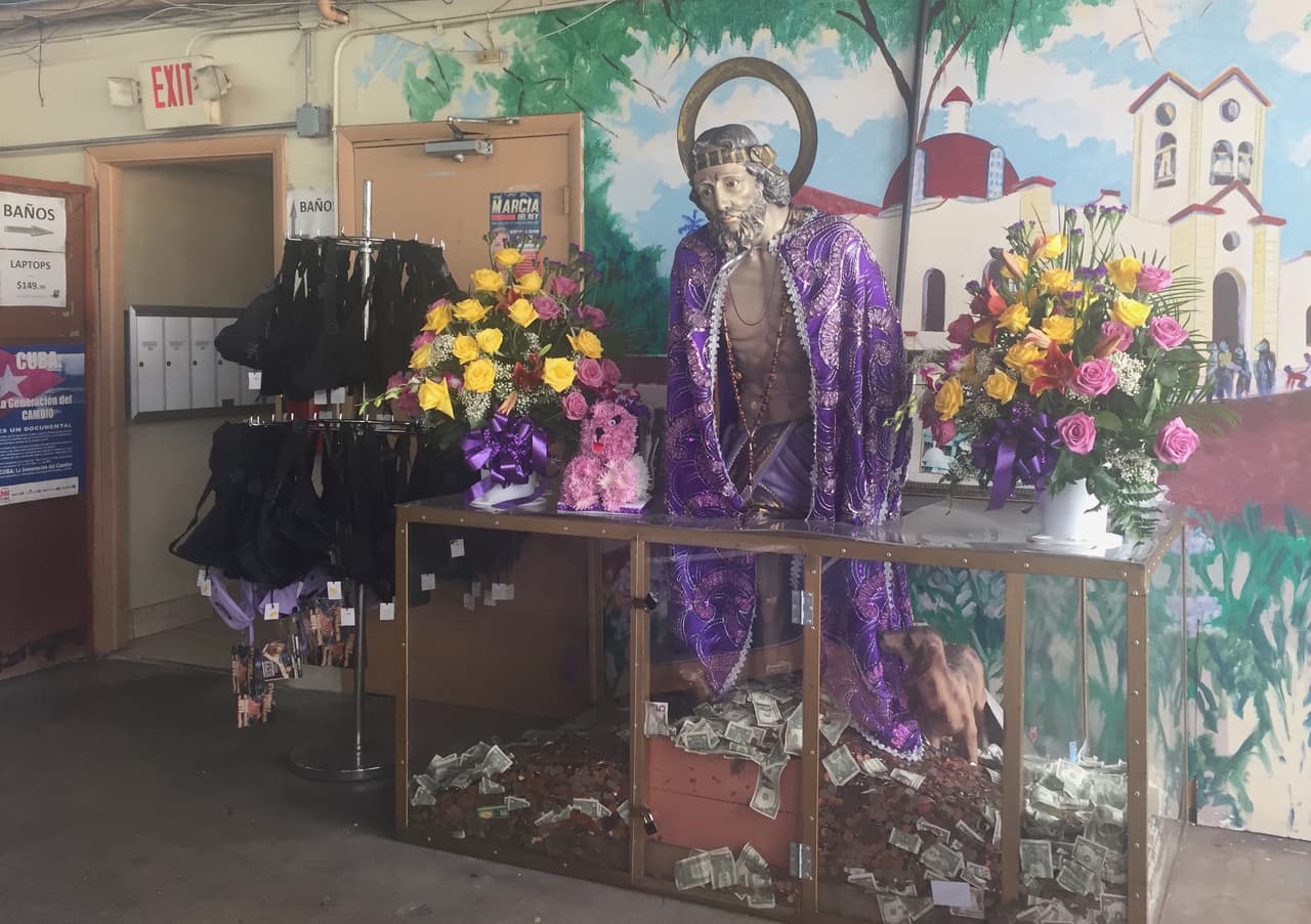 Al llegar a Ño que barato, de alguna manera se entra en Cuba. Tras subir por una rampa de madera, al comprador le recibe una imagen de San Lázaro, una figura especialmente venerada en Cuba por los santeros, con una capa púrpura y una ofrenda de dólares, y otra de San Judas Tadeo, el patrón de las causas difíciles.