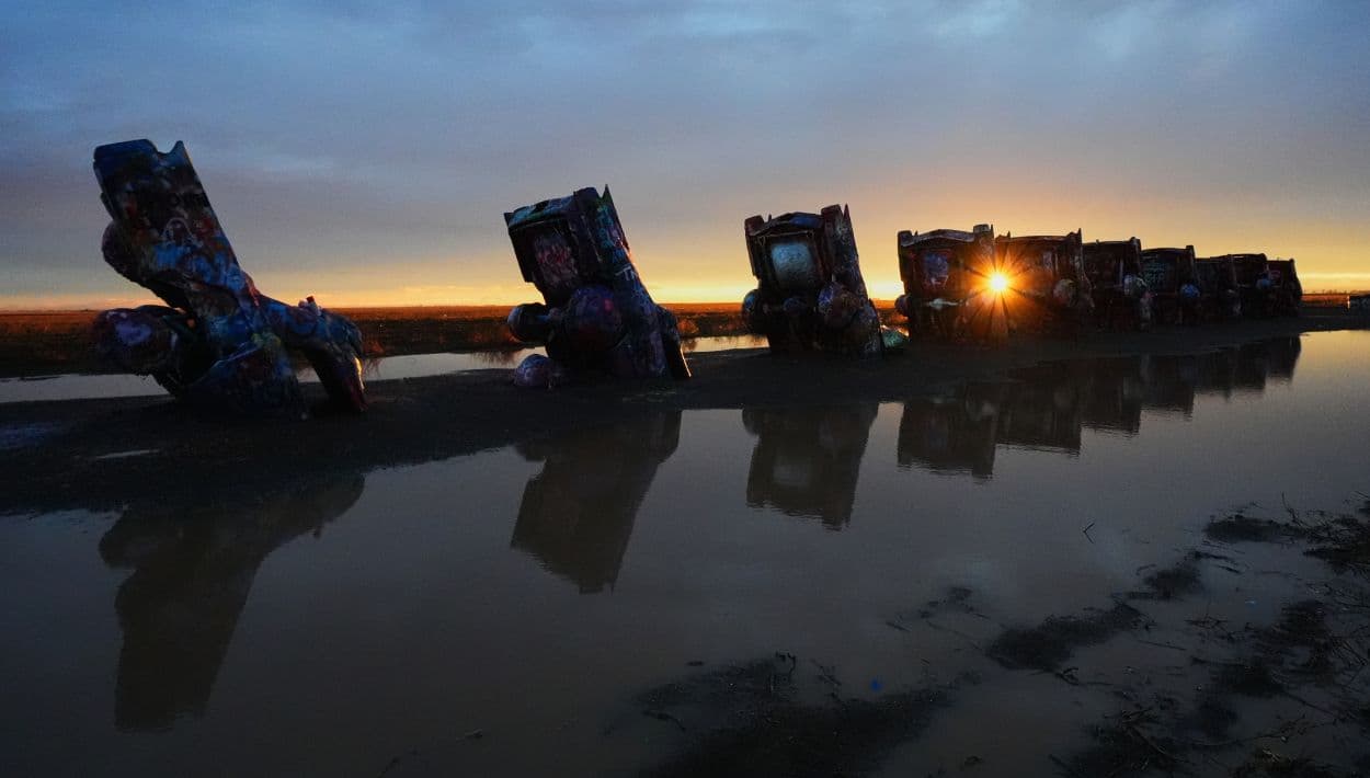 Un charco dejado por una tormenta del mediodía rodea vehículos mientras el sol poniente se asoma en Cadillac Ranch, una atracción al borde de la carretera a lo largo de la Ruta 66 en Amarillo, Texas.