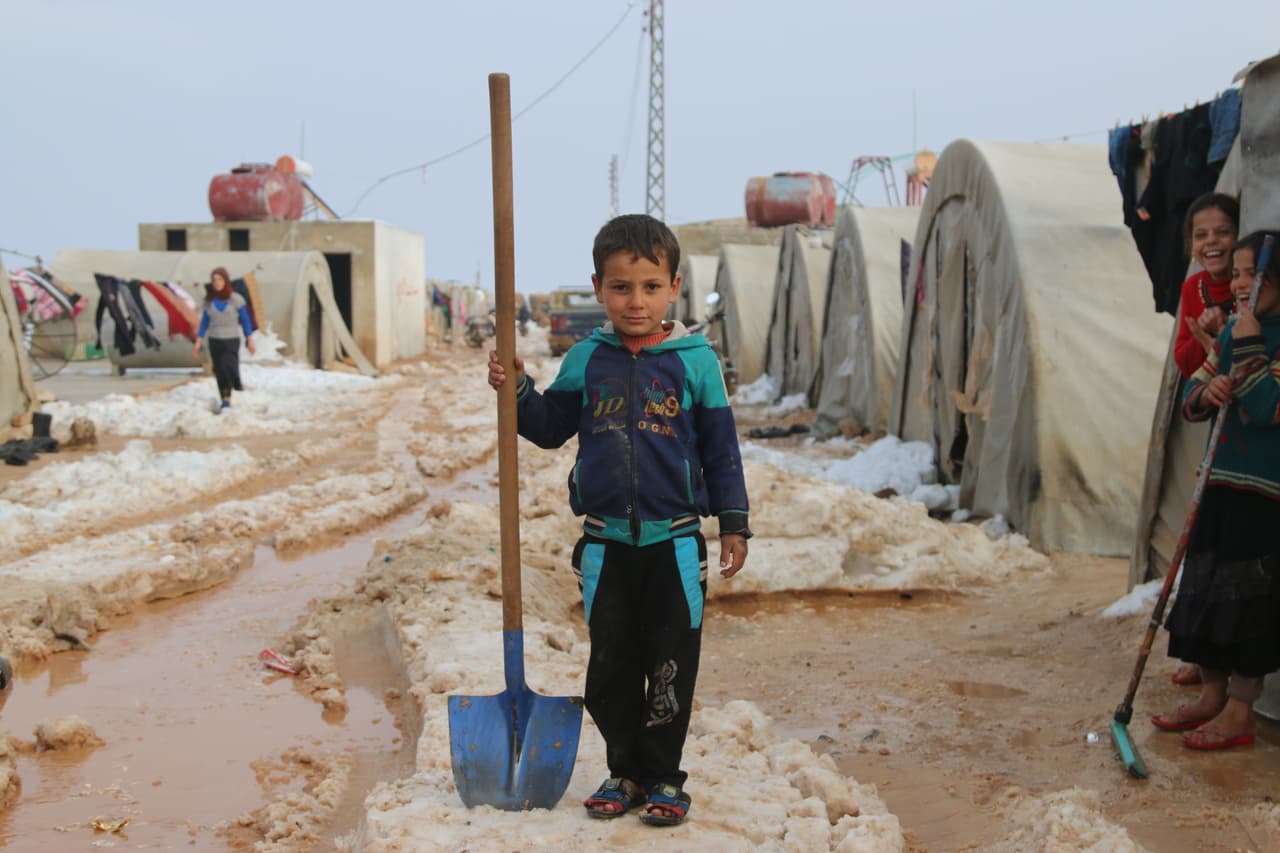 Radwan, de 6 años, frente a la carpa de su familia, en el campamento de desplazados de Al-Nour, Siria. Las dos hermanas de Radwan se ríen mientras él posa para la foto. Radwan y sus hermanas solo tienen zapatillas abiertas y las utilizan mientras despejan el hielo de la entrada de la tienda. "Si quitamos la nieve podemos dormir más calientes y mis pies no estarán fríos cuando salga", dice Radwan.