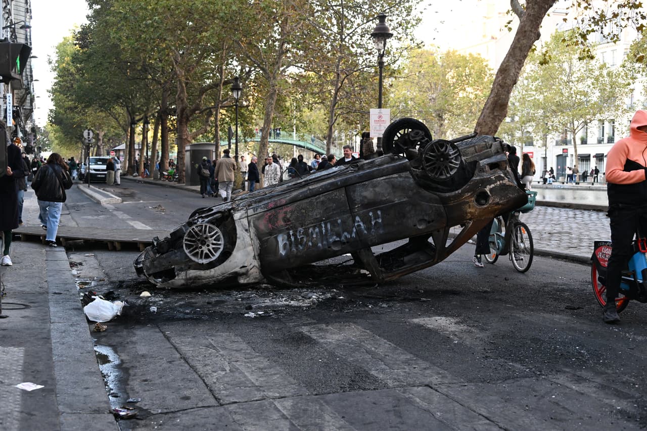 Varias personas pasan junto a un coche quemado al día siguiente de una manifestación contra el aumento del coste de la vida, sumándose a las quejas de los estudiantes, en París, el domingo 18 de octubre de 2026.