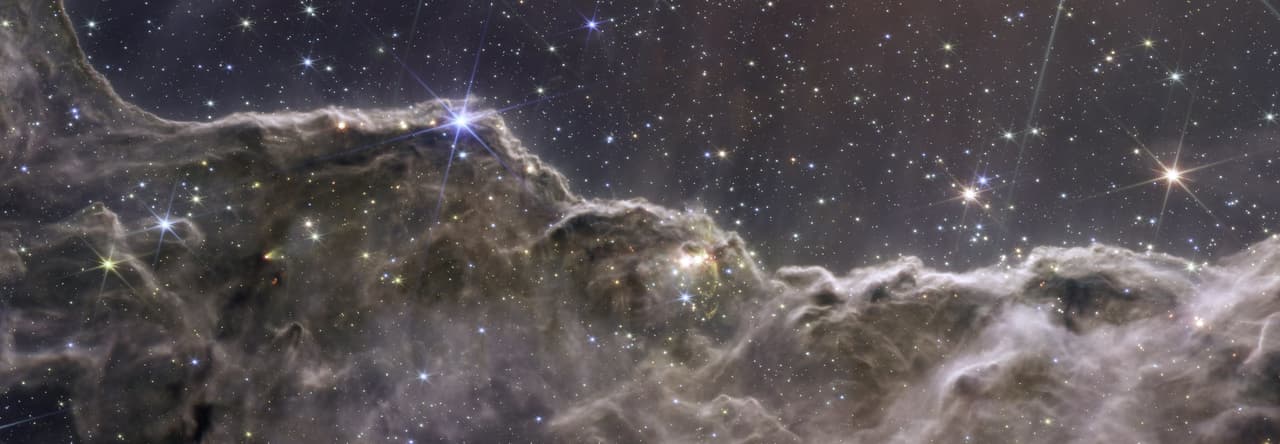¡Ha nacido una estrella! Nubes de gas y polvo de color marrón rosado dominan el primer plano de la imagen, brillando con estrellas jóvenes. Detrás de las nubes montañosas que brillan intensamente, el cielo parece azul marino, con estrellas y galaxias brillantes.