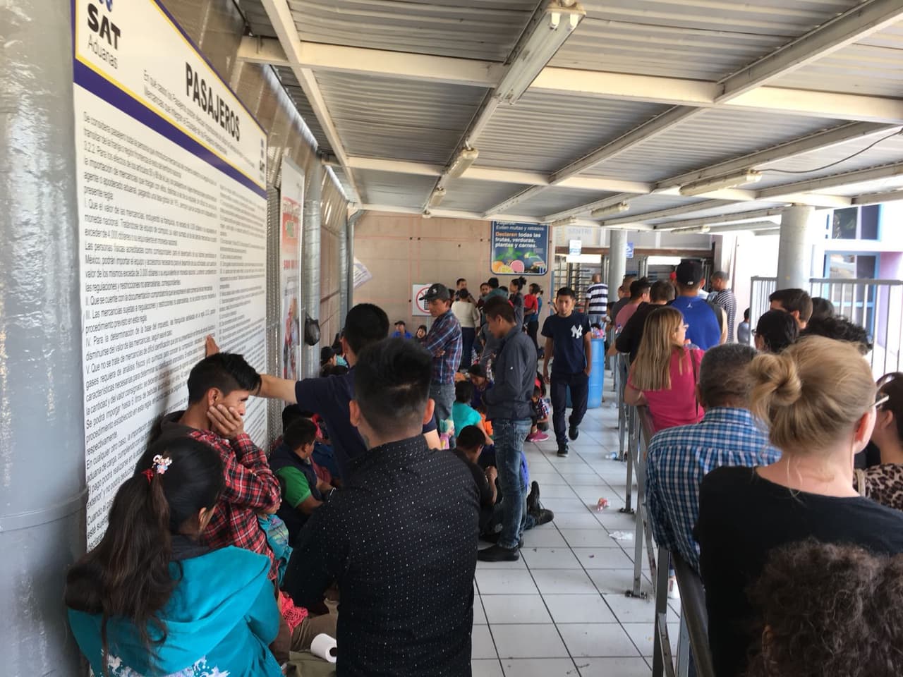 Los migrantes esperan sentados al lado de la fila de los transeuntes que esperan ser atendidos por los oficiales de aduanas.
