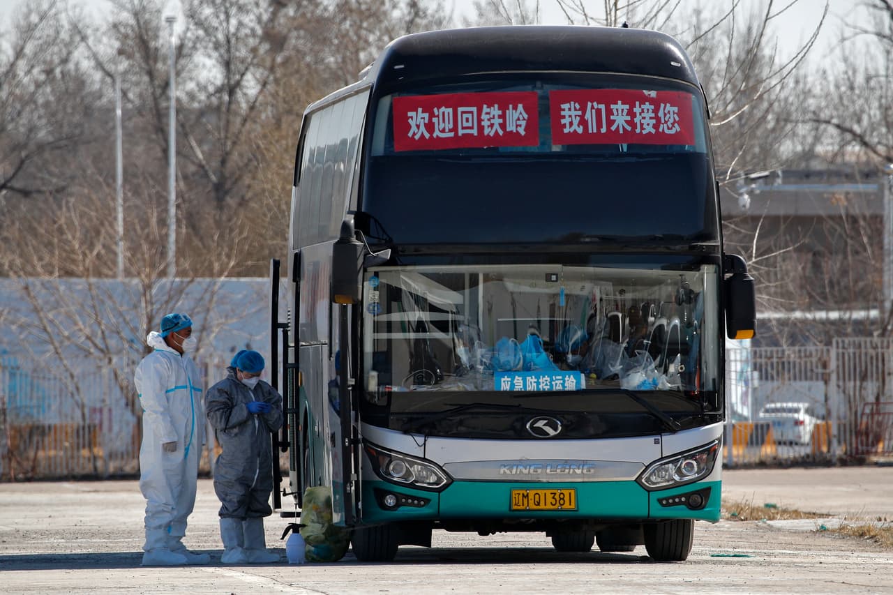 Los trabajadores que usan equipo de protección se paran afuera de un autobús utilizado para transferir a los viajeros a lugares de cuarentena fuera del nuevo Centro Internacional de Exposiciones de China convertido en un centro de tránsito para controlar las llegadas de ultramar a Beijing, el martes 17 de marzo de 2020.