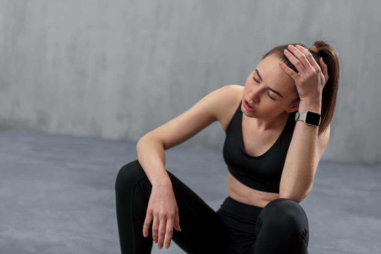 Tired young sporty woman having rest after workout, sitting on floor at gym , over grey background.