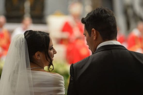 Esta imagen muestra a una pareja de novios en espera de ser unidos en matrimonio por el sacerdote que apenas se distingue sentado atrás.