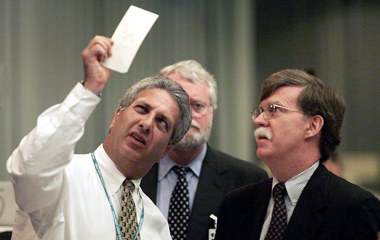 Un miembro de la junta electoral del condado de Palm Beach, en Florida, examina una papeleta junto al abogado republicano John Bolton (der.) y el abogado demócrata Gerry McDonough (centro) en el Centro de Operaciones de Emergencia de West Palm Beach en noviembre de 2000.