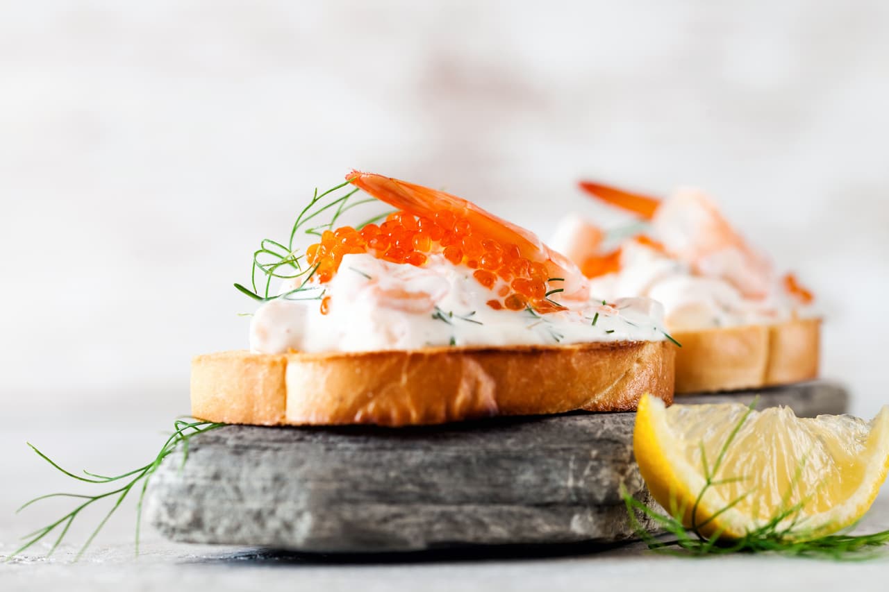 <b>Puesto 4. Toast Skagen</b>
<br>
<br>Es un sándwich sueco clásico y elegante de una sola cara que normalmente se sirve como aperitivo. Está hecho con pan blanco, camarones, mayonesa, mostaza de Dijon, huevas de pescado blanco, eneldo, limón y mantequilla. Se retiran las costras del pan y luego se saltea en mantequilla hasta que se doren.
<br>
<br>Los camarones se cortan y se mezclan con mayonesa, mostaza y eneldo. Luego se coloca la mezcla sobre el pan y se colocan las huevas encima de cada bocadillo. Se recomienda decorar cada tostada Skagen con una ramita de eneldo y servirla con una rodaja de limón al lado.
<br>
