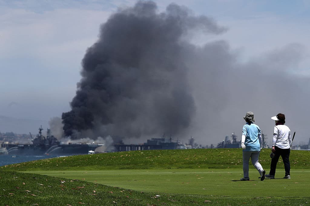 En la foto, golfistas caminando en un campo de golf mientras en el fondo se ve al USS Bonhomme Richard ardiendo.