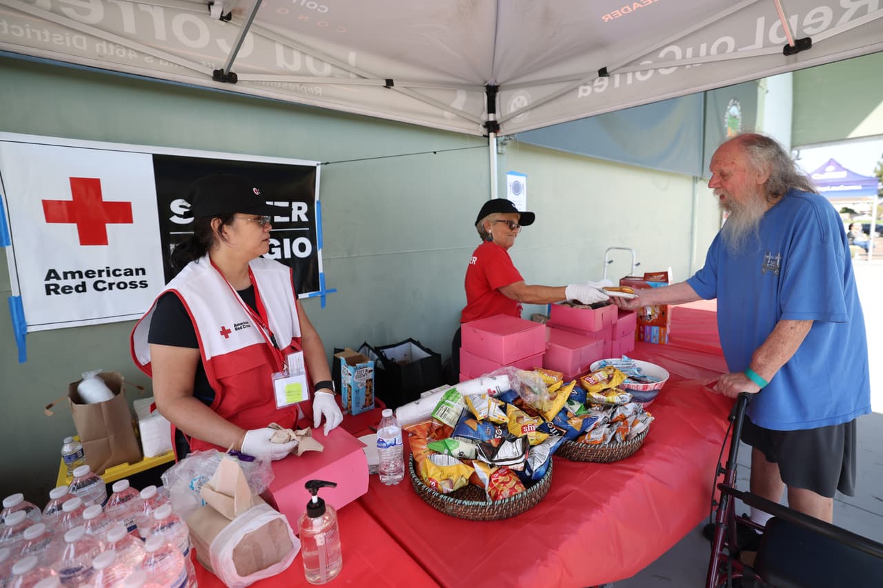 Las voluntarias de la Cruz Roja Americana, Florence Chong y Laura Siegelman, sirven refrigerios a Lenard Wilson, quien evacuó su casa, frente a Freedom Hall, un centro de evacuación en Fountain Valley, California, el sábado 23 de mayo de 2026. (Foto AP/Jill Connelly)