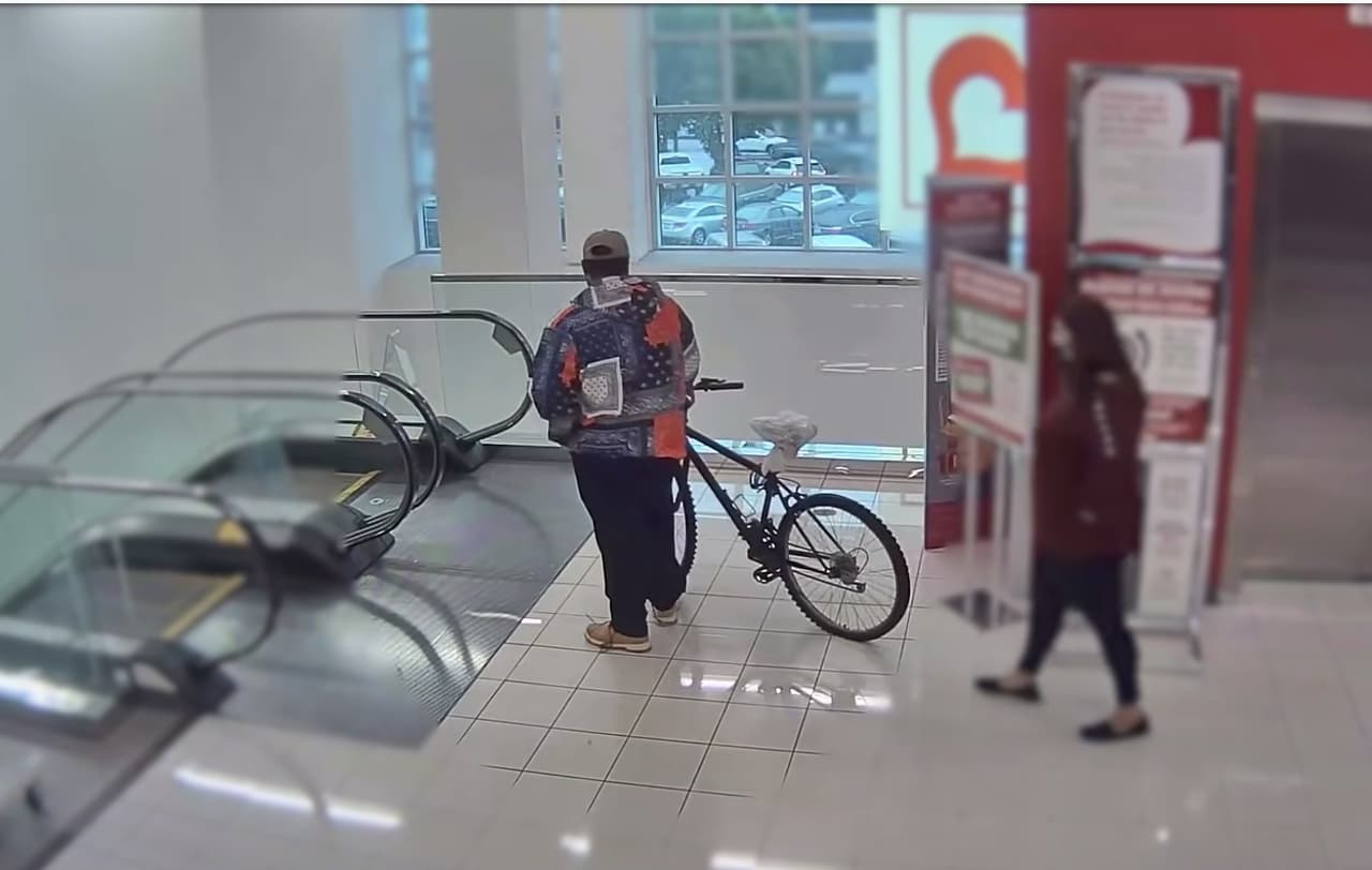 El hombre subió con la bicicleta al segundo piso de la tienda Burlington y comenzó a dar vueltas. Una mujer vestida con un saco rojo sigue los pasos del hombre que ve comportarse de modo sospechoso.