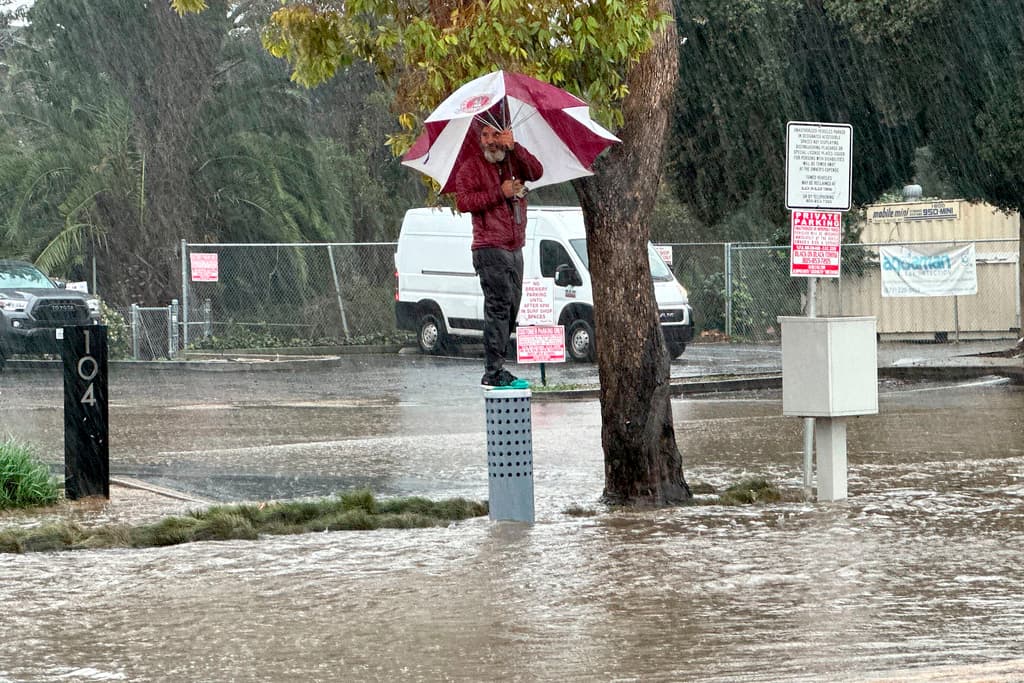 Un hombre que lleva un paraguas se encuentra sobre una calle inundada en Ventura, California, el domingo 4 de febrero de 2024. (Foto AP/Eugene García)