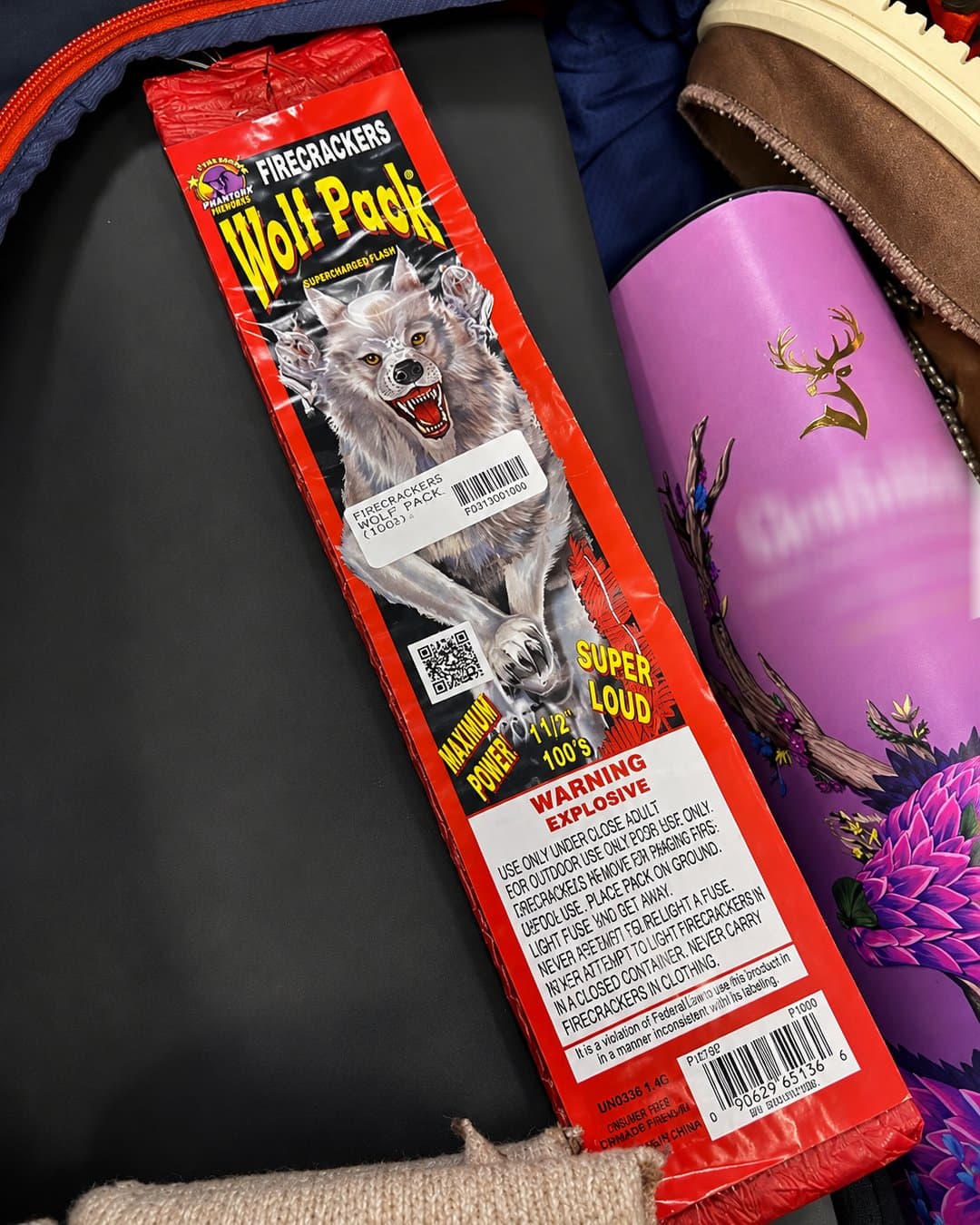 Paquete rojo de petardos "Wolf Pack" con la imagen de un lobo furioso, advertencias de explosivos en inglés, códigos de barras y un cilindro rosado decorado con flores a un lado.