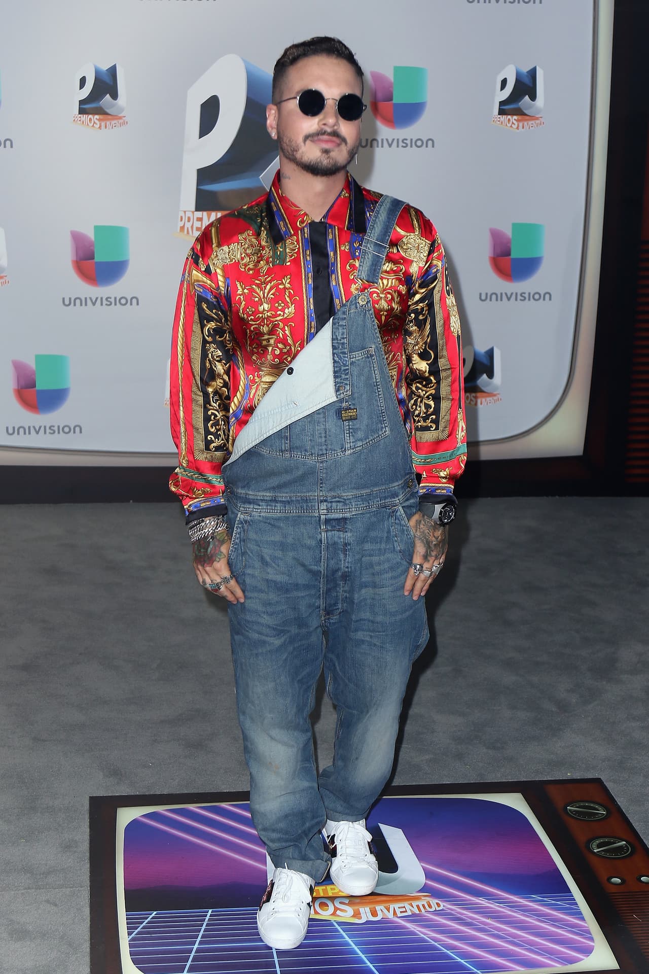 J Balvin se fue en busca de referencias vintage y se topó con las emblemáticas camisas de de cadenas de Versace propias de los noventa. para serle fiel a su estilo, la combinó con un overol de jean y unos elegantes tenis Gucci.