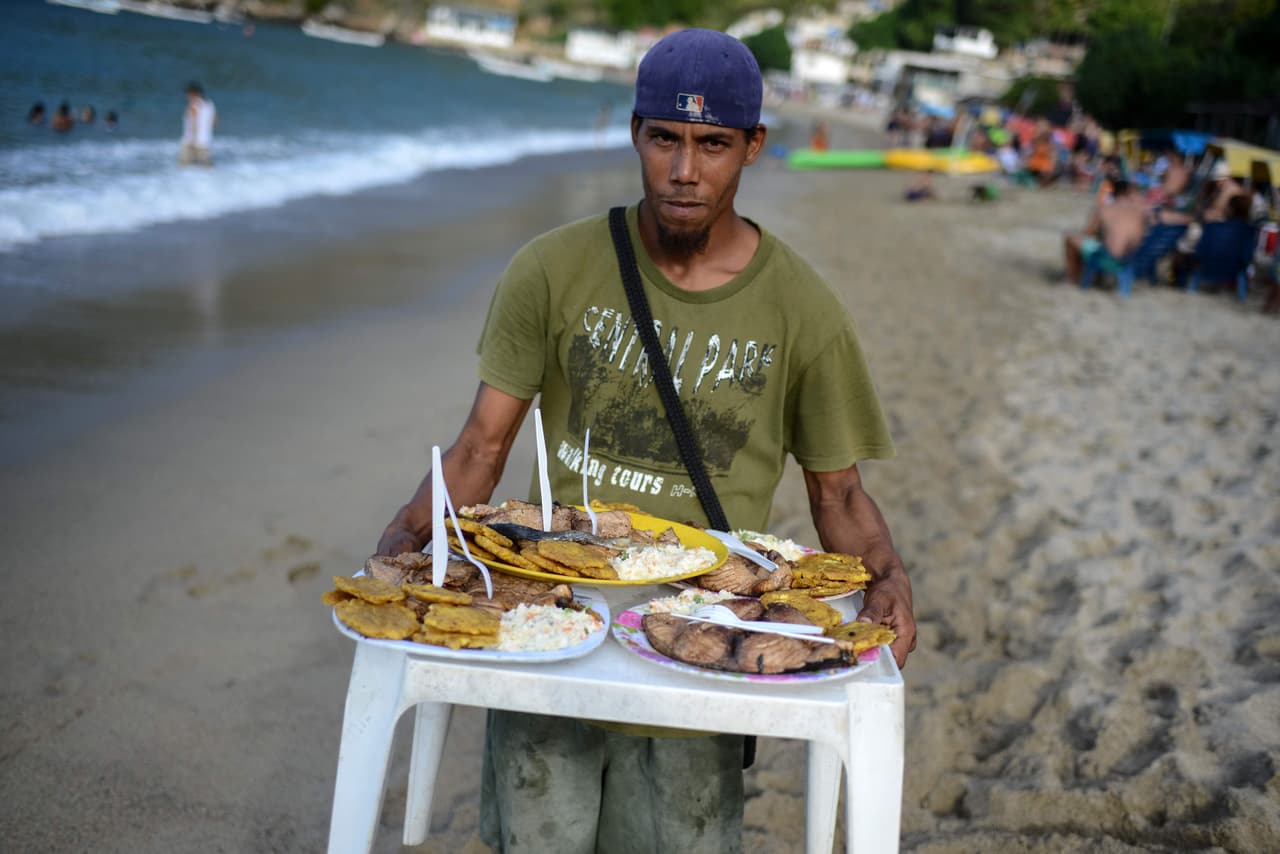 Un empleado de un restaurante a la orilla del mar lleva los tradicionales platillos de pescado frito con plátano a los comensales. Frangeli Mayora, propietaria de un negocio de alquiler de sillas de playa y madre de dos niños, lamenta que casi nadie trae efectivo para pagar sus servicios. “Es arrecho (duro). Desde que (el expresidente Hugo) Chávez murió, esto es una locura”, afirmó la comerciante.