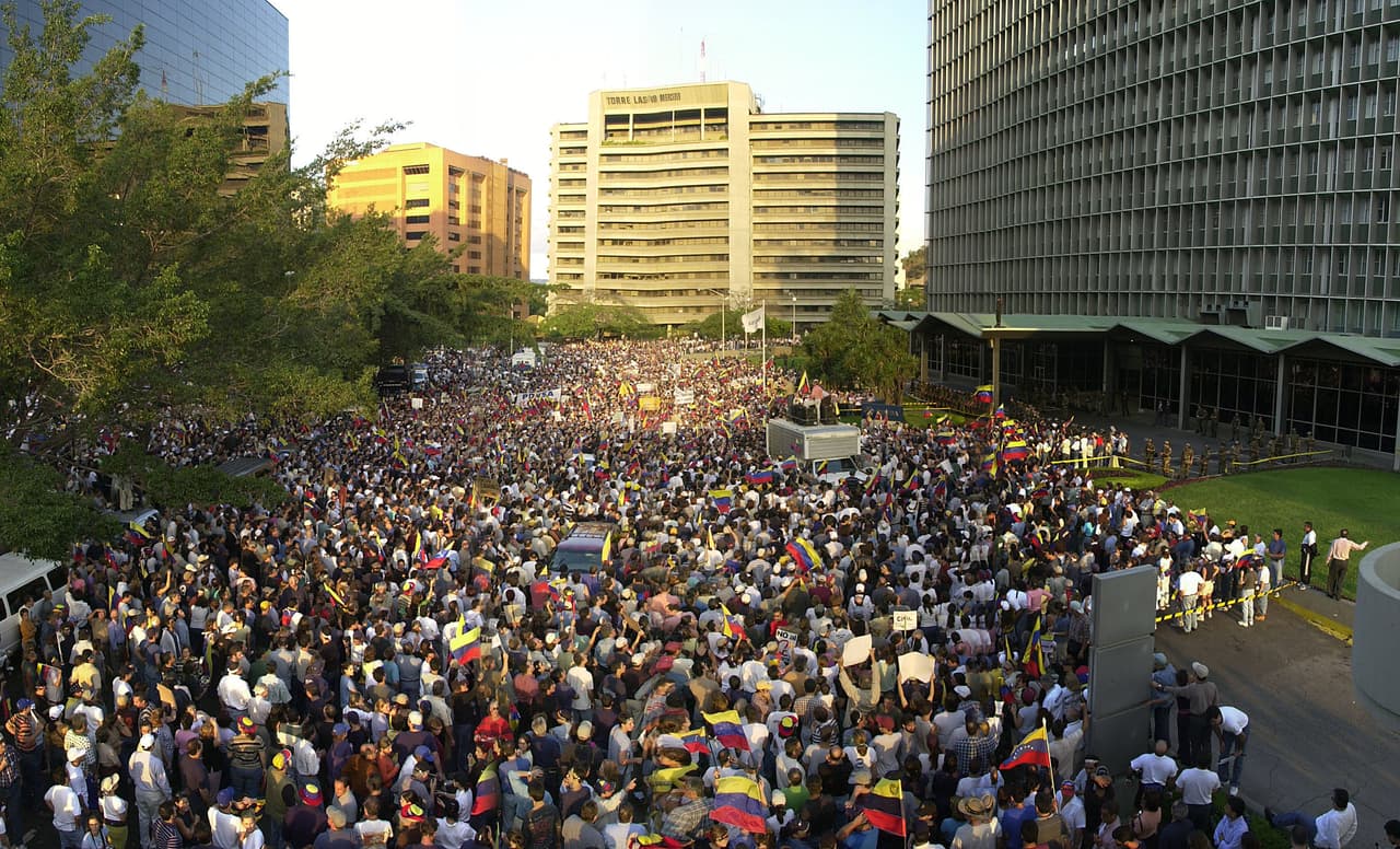 <b>Oleada de protestas.</b> Al comienzo de su nuevo período presidencial en 2001, Hugo Chávez fue habilitado por el Parlamento de mayoría chavista para gobernar por decreto, lo que le permitió hacer un paquete de leyes unilateralmente. Eso desató una ola de protestas civiles y algunos militares activos se manifestaron contra el presidente. Los partidos de oposición presionaban, también la principal organización empresarial (Fedecámaras) y el sindicato nacional de trabajadores (CTV). El 7 de abril de 2002, Chávez despidió en televisión nacional a los más altos gerentes de la petrolera estatal PDVSA, que reclamaban la nueva designación de directivos de la empresa (prochavistas) que no cumplían los méritos para ocupar esos cargos. En la fotografía, tomada el 9 de abril, los manifestantes se reúnen frente a la sede de PDVSA para protestar los despidos.
