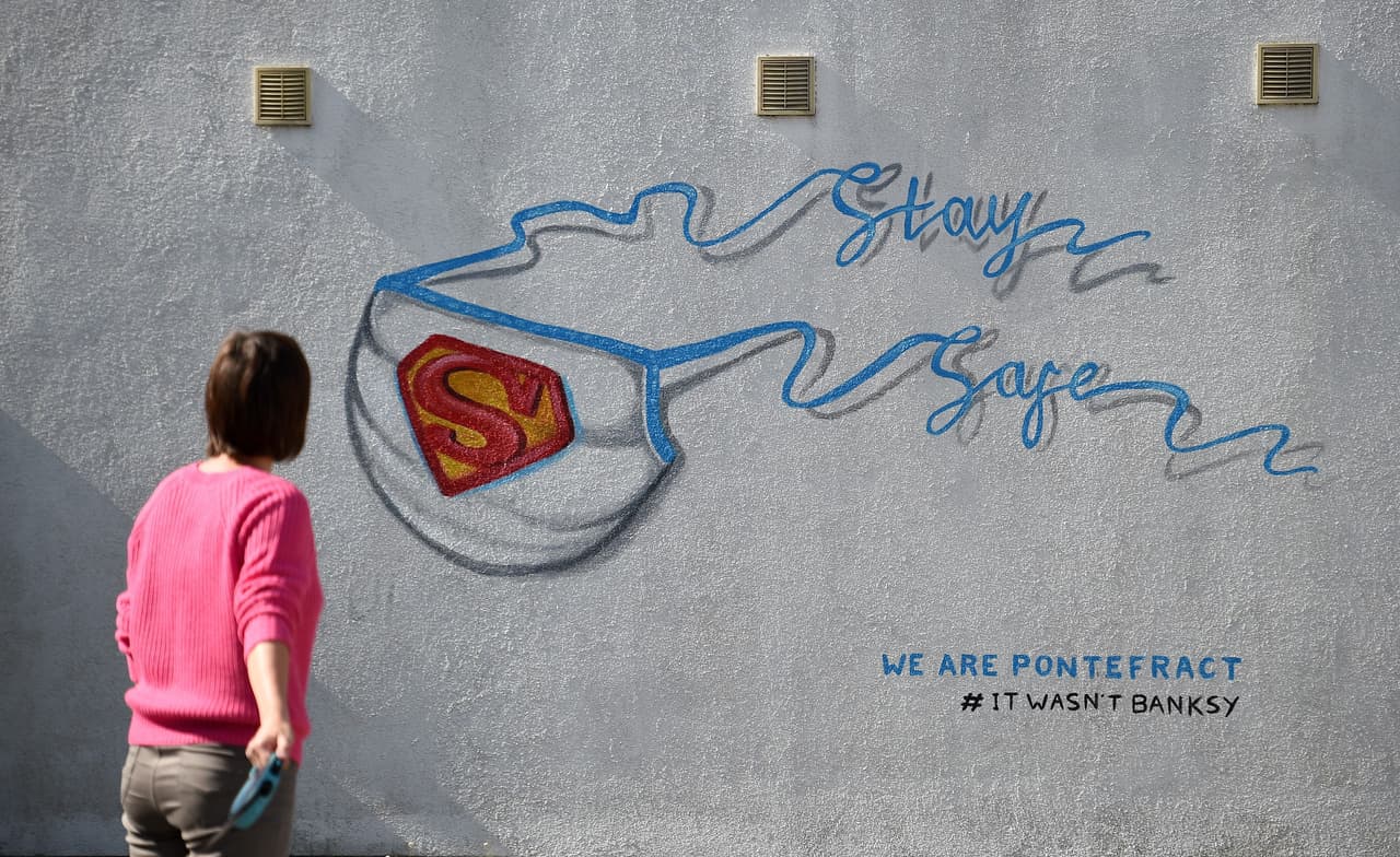 Otro grafiti que combina la iconografía de uno de los más famosos superhéroes (Superman) con elementos relacionados con el personal sanitario y la atención médica. En la imagen, una mujer que pasea a su perro se detiene a mirar este grafiti en una pared de Pontefract, al norte de Inglaterra. El logo de Superman aparece aquí sobre una mascarilla cuyos elásticos conforman la expresión "Quédense a salvo".