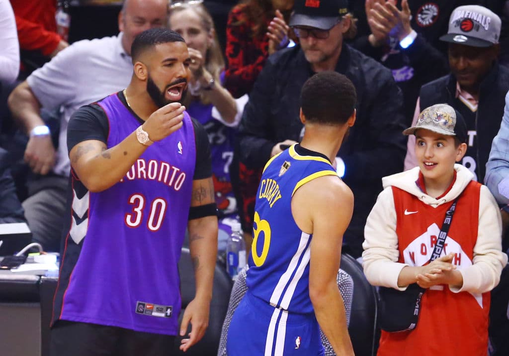 El rapero Drake tratando de meterse en la cabeza de Stephen Curry mientras un joven aficionado observa y escucha el díalogo.