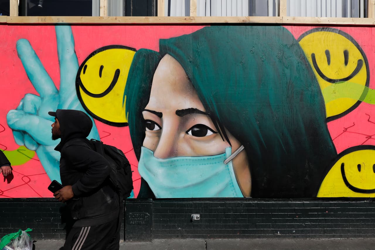 Una mujer con mascarilla y guantes haciendo el símbolo de la paz con las manos y rodeada de pequeñas caritas sonrientes estilo emoticon, adorna la fachada de un comercio cerrado en el centro de Seattle. Se trata de una obra que busca dar aliento durante esta tragedia, hecha por los artistas @theydrift y @ksra_ksra.