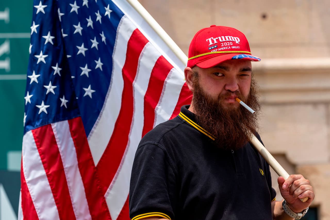 Aquí uno de los manifestantes sin mascarilla, para protegerse del coronavirus, carga una bandera de EEUU y lleva puesta una gorra de Trump 2020.
<br>