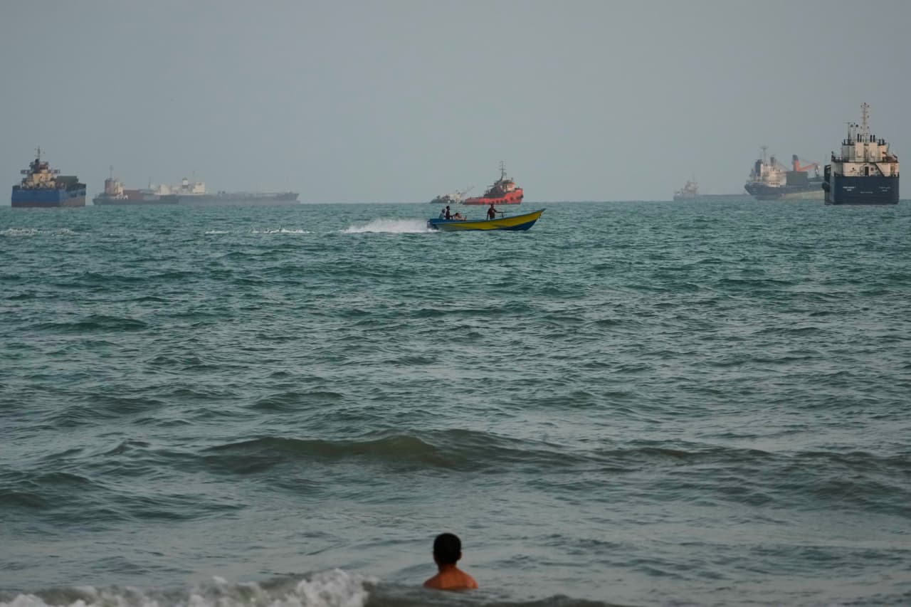 Un nadador permanece cerca de la costa al tiempo que una lancha a motor pasa junto a buques comerciales anclados en el estrecho de Ormuz