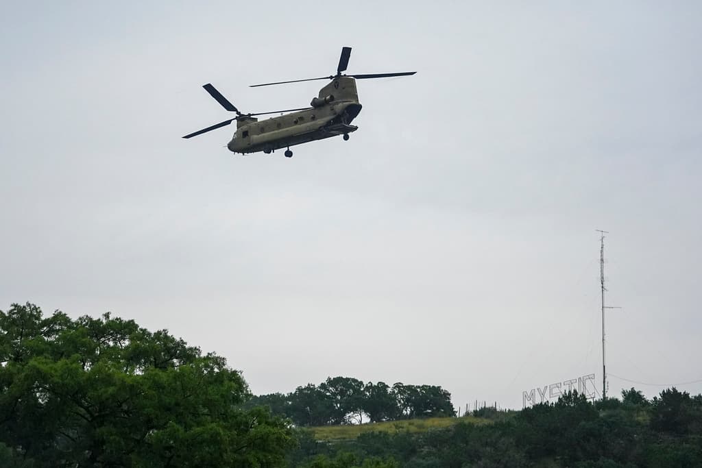 Un letrero de Camp Mystic se ve sobre una colina, mientras un helicóptero sobrevuela el río Guadalupe en busca de sobrevivientes o víctimas.