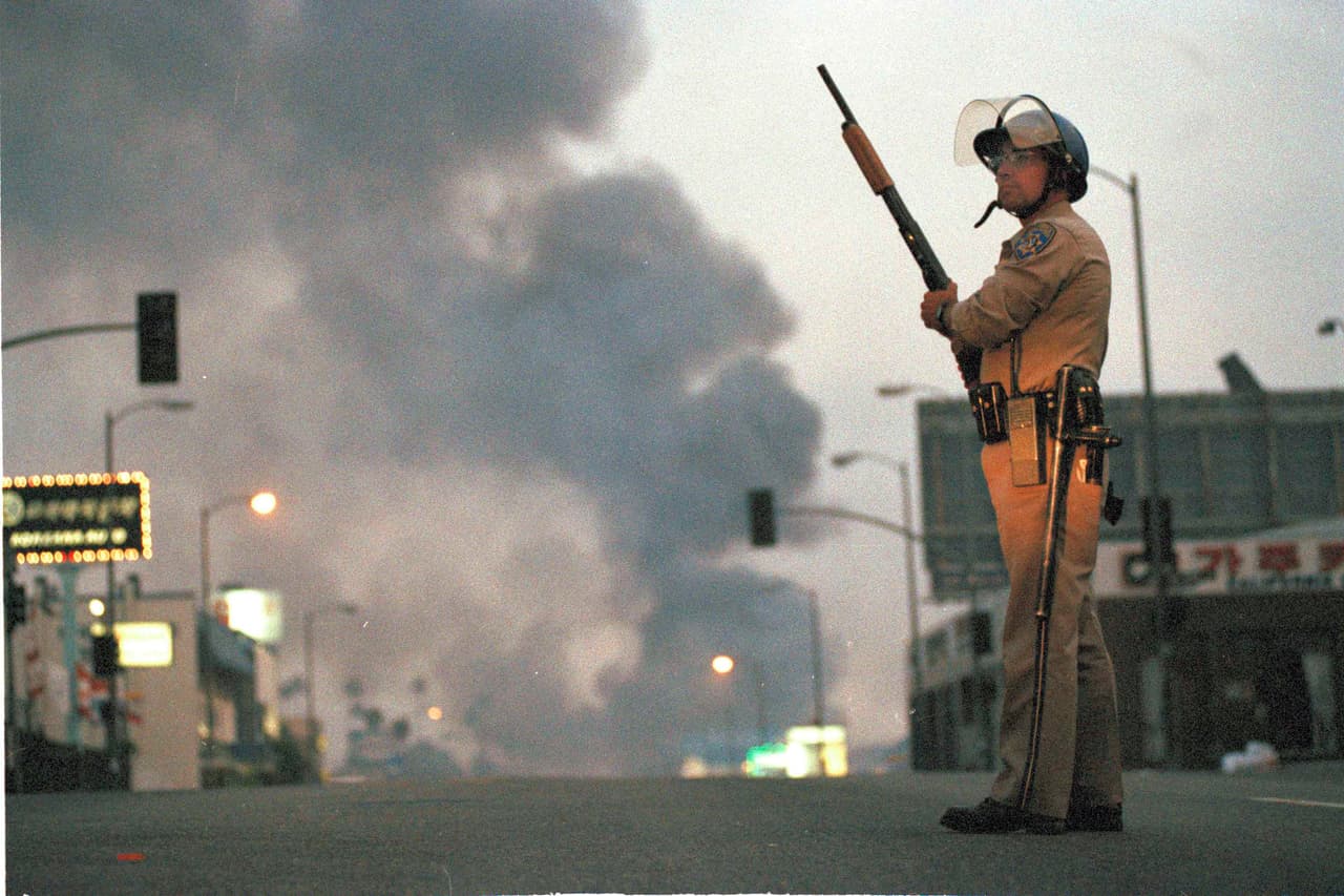 Un agente de la Patrulla de Carreteras de California vigila la esquina de las calles 9 y Vermont en Los Ángeles, mientras se observa de fondo una cortina de humo por un incendio. Era el 30 de abril de 1992, el segundo día de disturbios en la ciudad.