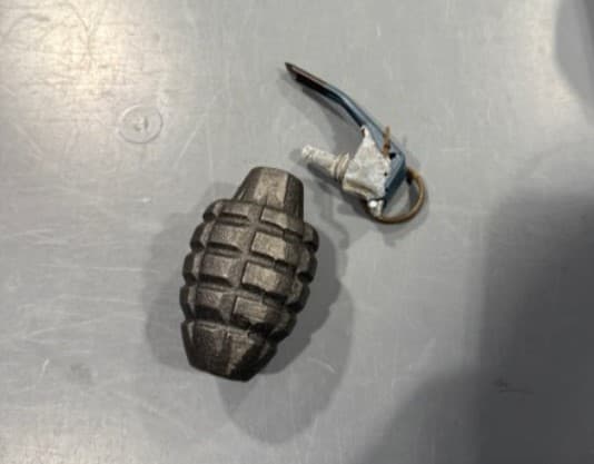 Granada de mano de práctica Mk 2 desarmada sobre una superficie gris de la TSA. El cuerpo metálico texturizado está separado de la espoleta, el pasador de seguridad y el anillo de tracción. Objeto no funcional, pero prohibido en vuelos.