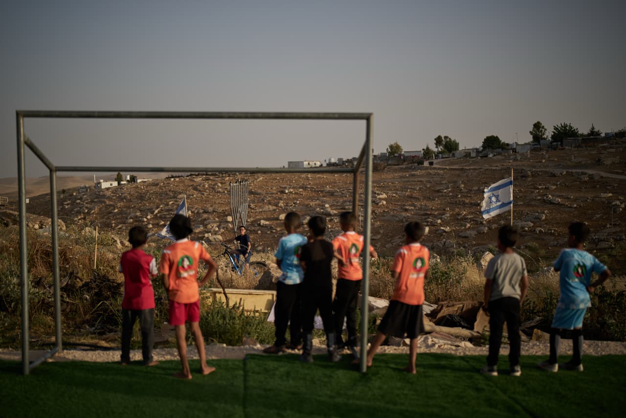 Unos niños palestinos miran a un joven colono israelí después de que les quitaran su balón de fútbol, que había sido pateado más allá de una cerca de alambre de púas que rodeaba un pequeño campo de fútbol cerca del asentamiento de Carmel, en la aldea beduina de Umm al-Khair, en Cisjordania, el martes 12 de mayo de 2026. (Foto AP/Leo Correa)