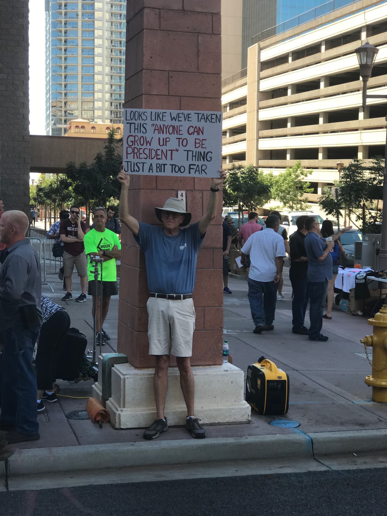 Este hombre porta un cartel que dice que dice: "Cualquiera puede crecer y ser presidente" mientras se mantiene en la sombra a las afueras del Centro de Convenciones.