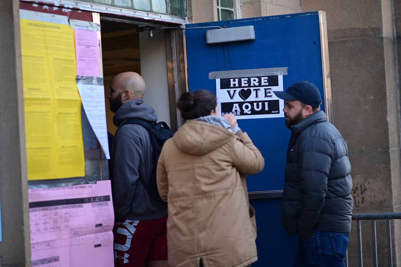 Un cartel dice "AQUÍ VOTE AQUI" mientras los votantes esperan en la fila mientras el centro de votación del Centro Recreativo Kendrick abre a las 7 a. m. el 8 de noviembre de 2022 en Filadelfia.