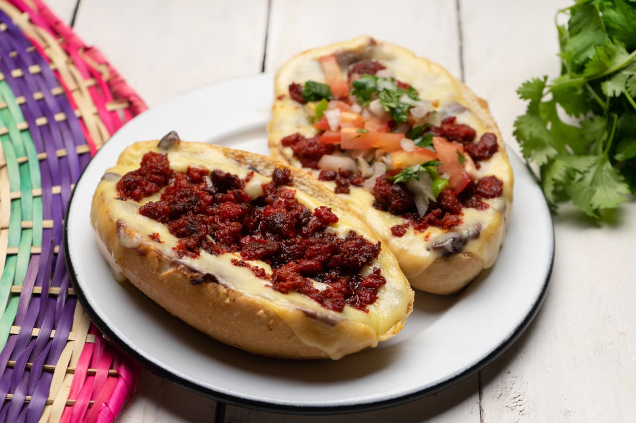 <b>Puesto 14. Mollete </b>
<br>
<br>Este sándwich tradicional del norte de México consiste en la mitad de un pan bolillo cubierto con frijoles refritos, queso y salsa de tomate. Se puede encontrar en todo México en numerosas cafeterías, restaurantes y puestos de comida callejera.
<br>
<br>El sándwich tiene raíces en Andalucía, España, donde se llama molletes a un pan rústico ovalado que ha sido un alimento básico en el desayuno durante muchos años. Sin embargo, la versión española es mucho más simple, solo lleva aceite de oliva, tomate y ajo.
<br>
<br>Los molletes mexicanos se consideran primos lejanos de la bruschetta italiana y al sur del país a menudo se sirven con pico de gallo, tocino y champiñones.
<br>