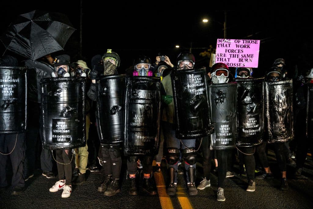 Muchos manifestantes vestidos de negro, armados con escudos, cascos, y máscaras antigases, ocupan la primera línea, preparados para la carga policial.