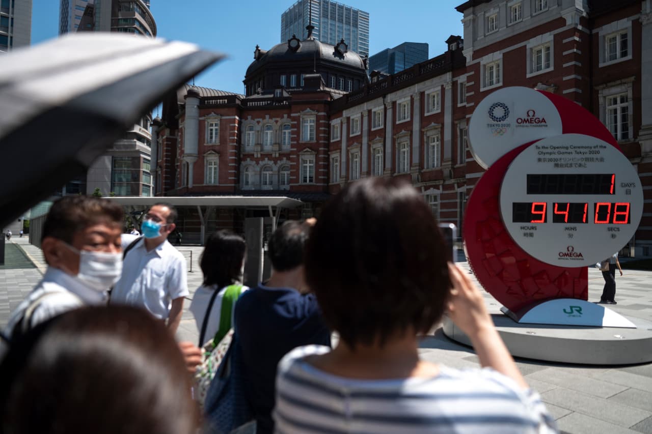 Protegidos con mascarillas a pesar de estar al aire libre, un grupo de personas toma fotografías del reloj que indica la cuenta regresiva para el comienzo oficial de los juegos, un día y nueve horas antes del inicio de la ceremonia de apertura.