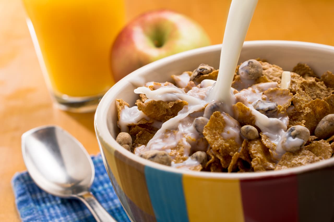 A breakfast still life consisting of a bowl of cereal, pouring milk, a glass of orange juice, an apple, and a spoon.