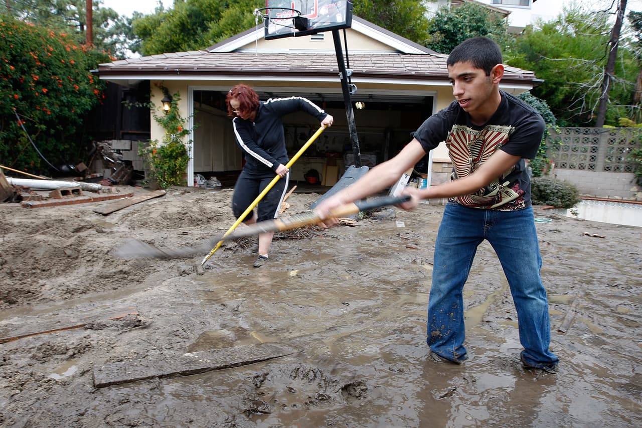 LA CANADA-FLINTRIDGE, CA - FEBRUARY 6: Matthew Fernandez helps a neighbor shovel ankle-deep mud after debris flows damaged homes and carried cars away following heavy rains that caused mudslides on February 6, 2010 in La Canada Flintridge, California. Large wildfires in 2008 and 2009 stripped the hills and mountains of vegetation, resulting in mud and debris flow danger as winter rains pass over foothill communities where thousands of people have been evacuated at times in recent weeks. The threat is particularly high near the San Gabriel Mountains above La Canada-Flintridge area which were denuded of natural flood-controlling vegetation by the 250-plus square mile Station. At least 40 homes have been severely damaged and 500 remain evacuated. (Photo by David McNew/Getty Images)