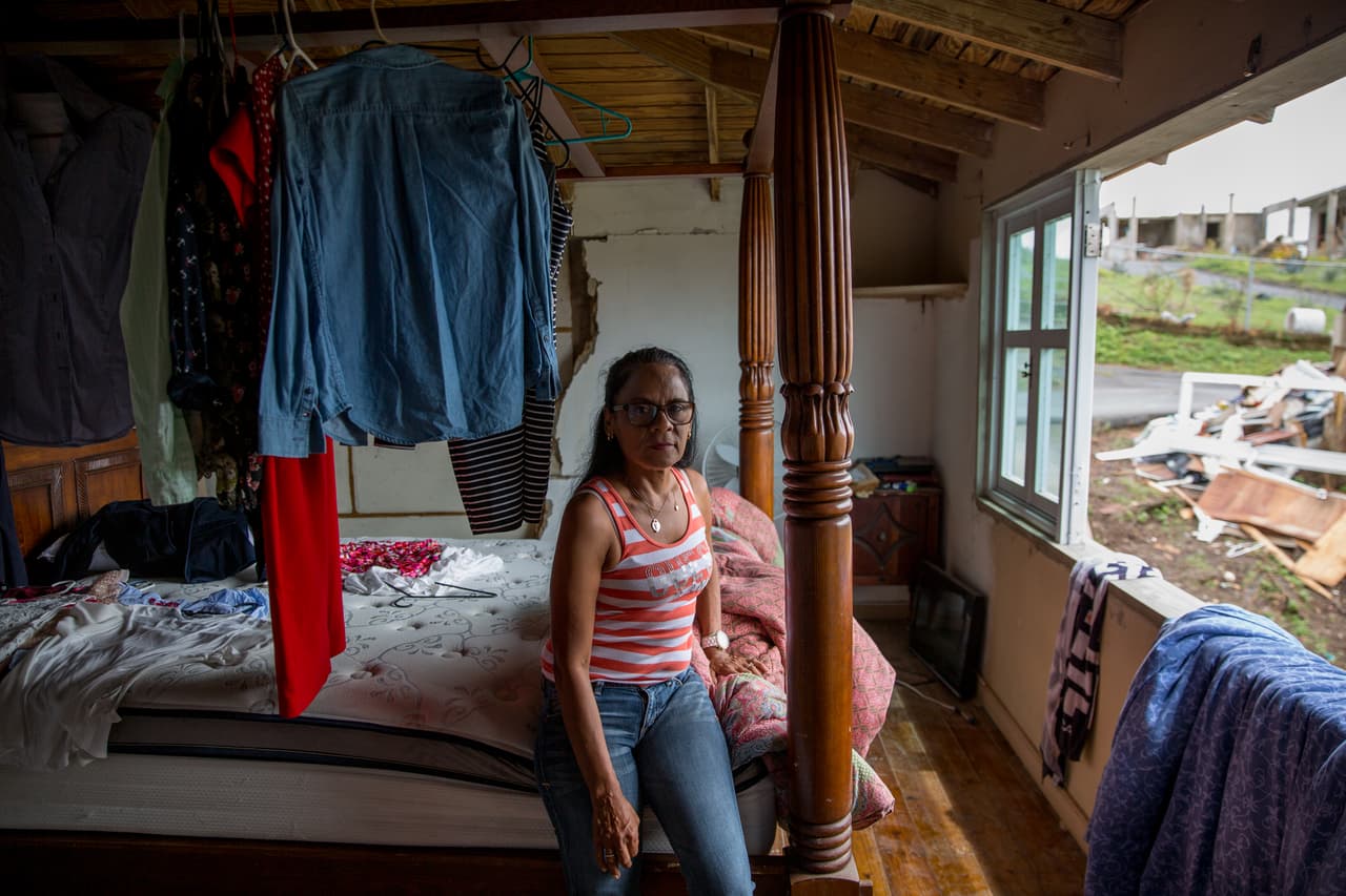 Carmen Virginia Delerme posa en lo quedó de su recámara, destruida por el techo de su vecino que se desprendió por la fuerza de María. Delerme pasó la peor parte del huracán junto a su hija en la escuela que dirige. Al día siguiente no podía entender camino a casa por qué su vecino se disculpaba. Luego descubrió que su hogar debía ser reconstruido.