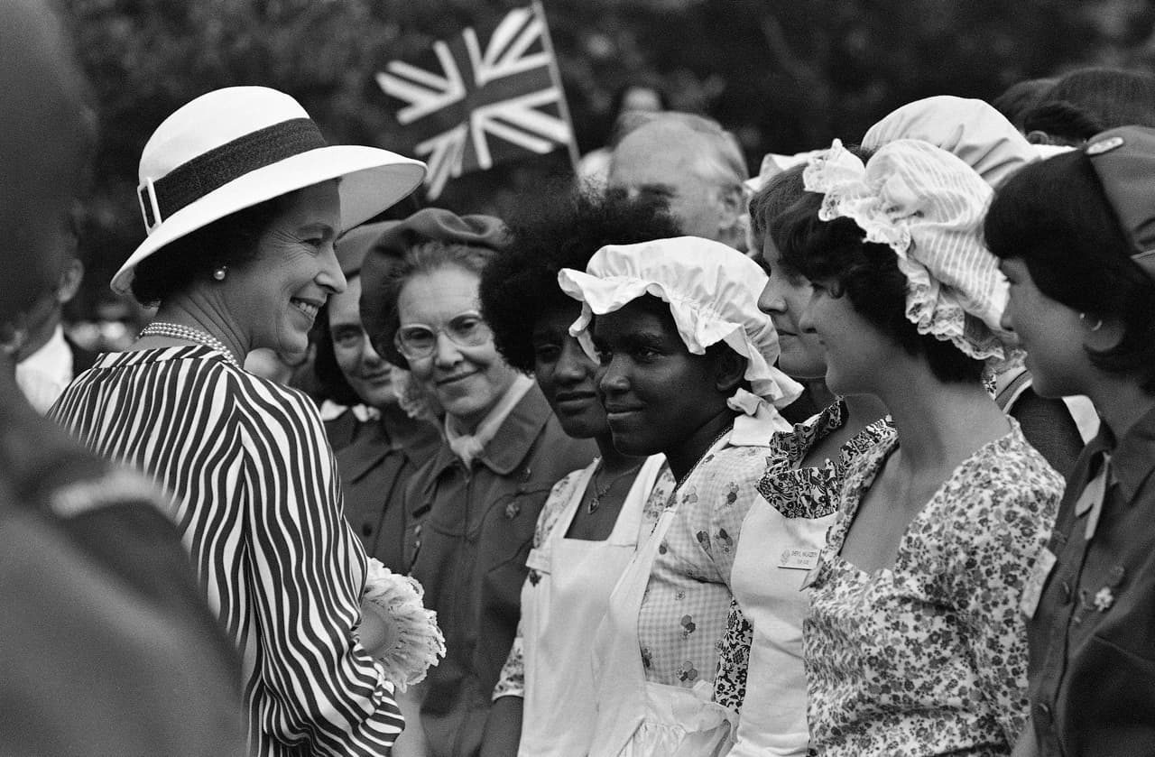 La reina Isabel II sonríe a las Girl Scouts, vestidas con atuendos coloniales, mientras las Girl Scouts of America le presentan un libro sobre la mujer estadounidense a la Reina.