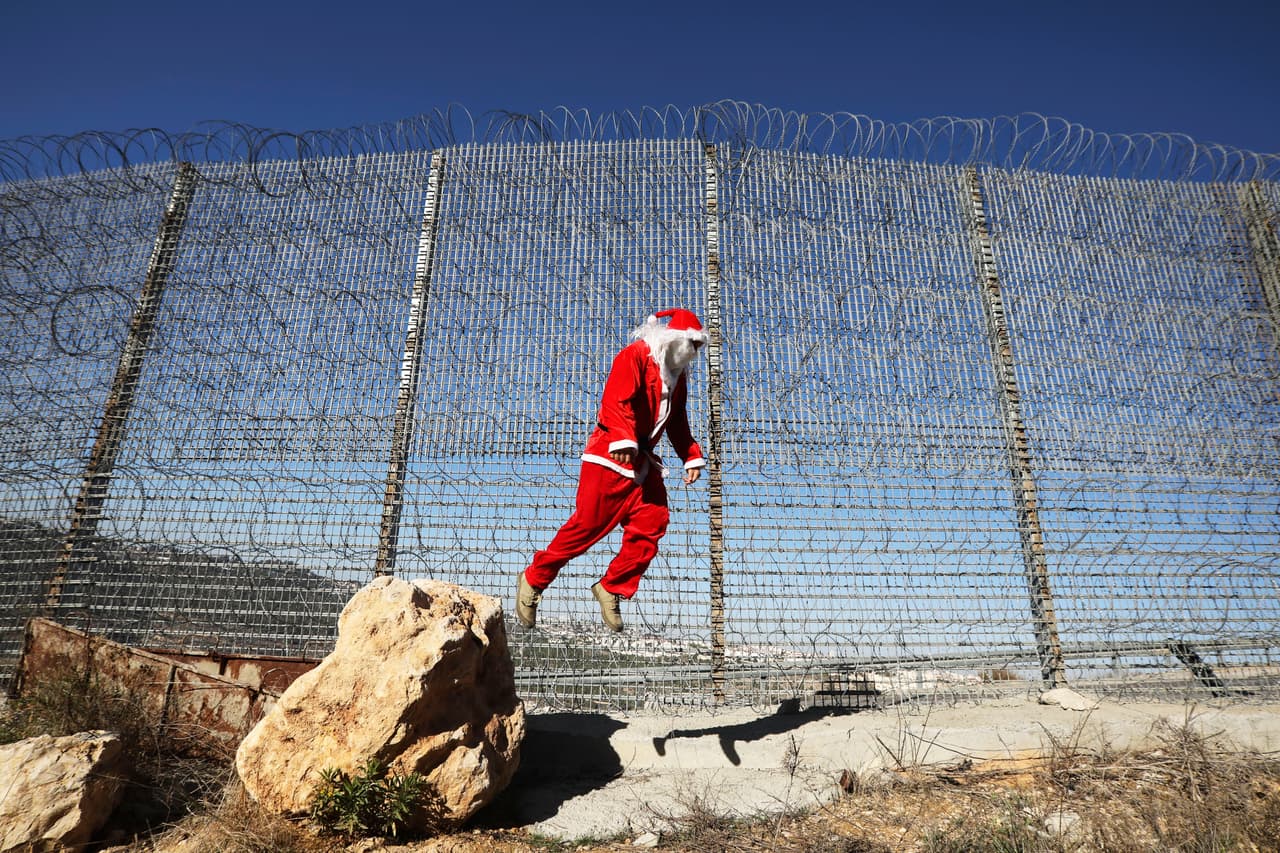 Un joven palestino disfrazado de Santa Claus salta junto a la valla fronteriza con Israel, durante un protesta en la ciudad de Belén. 23 de diciembre de 2019.