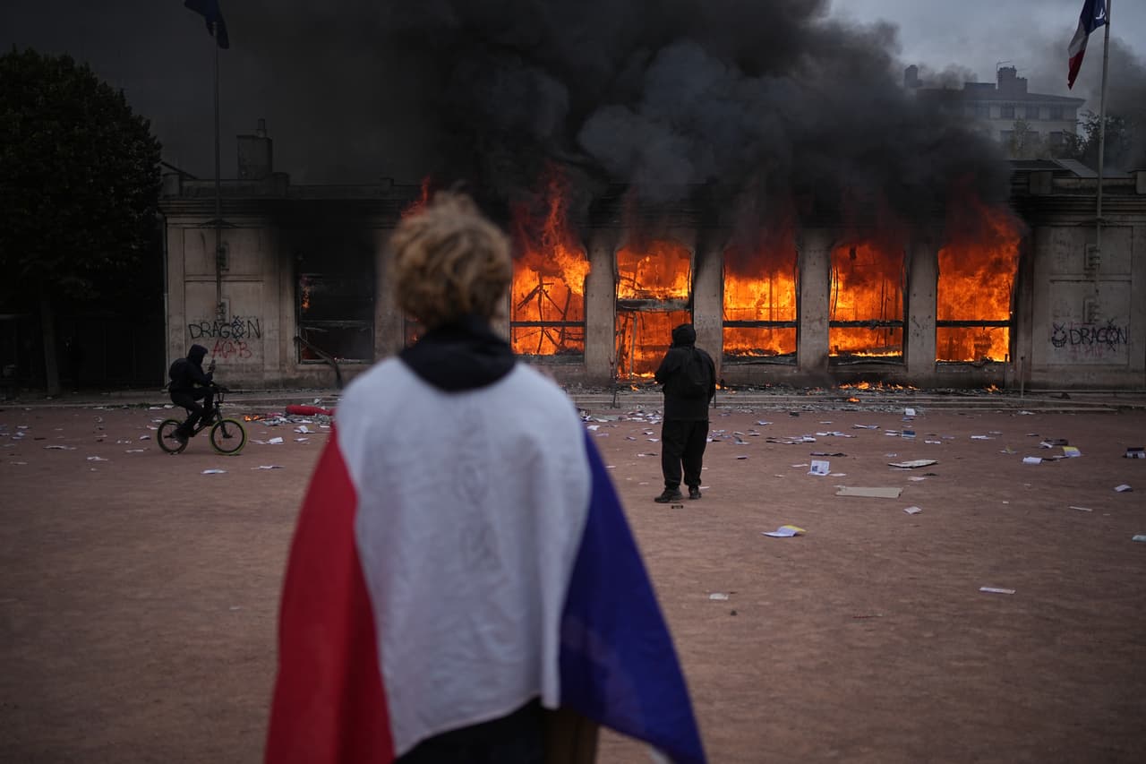 Un manifestante se envuelve en una bandera francesa frente a la oficina de turismo en llamas durante una protesta contra el aumento del costo de vida y en solidaridad con las reivindicaciones estudiantiles en Lyon, Francia, el sábado 17 de octubre de 2026.