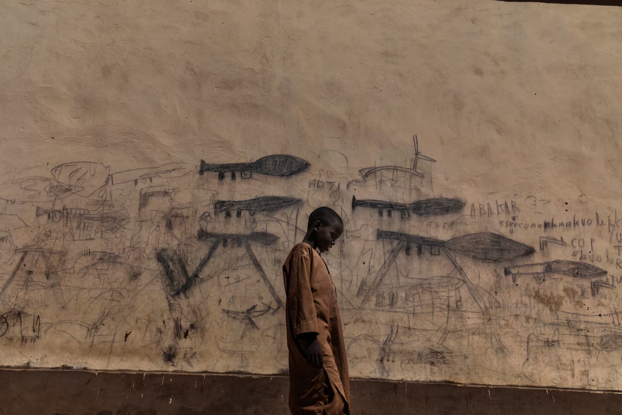Esta imagen nominada a fotografía del año muestra a un niño que camina junto a una pared con dibujos de armas y explosivos en Bol, Chad, el 16 de octubre de 2018. Una crisis humanitaria está en curso en la cuenca del lago Chad, causado por una compleja combinación de conflicto político y factores ambientales. Este lago fue uno de más grandes de África pero se ha ido secando y ha dejado sin medios de subsistencia a muchas personas. El grupo yihadista Boko Haram, que está activo en el área, se beneficia de las dificultades y utiliza las aldeas locales como lugares de reclutamiento de jóvenes.