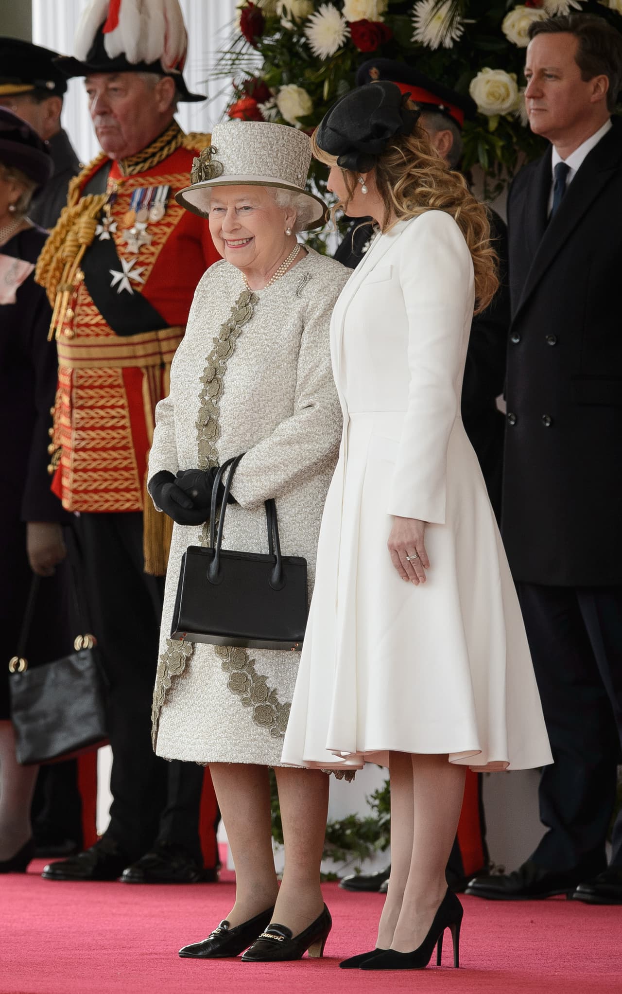 Esa pieza tradicional la combinó con un vestido tipo abrigo blanco y complementó su look con una bolsa tipo clutch y stilettos negros. La reina optó por tonos dorados con detalles en verde oliva en su vestimenta.