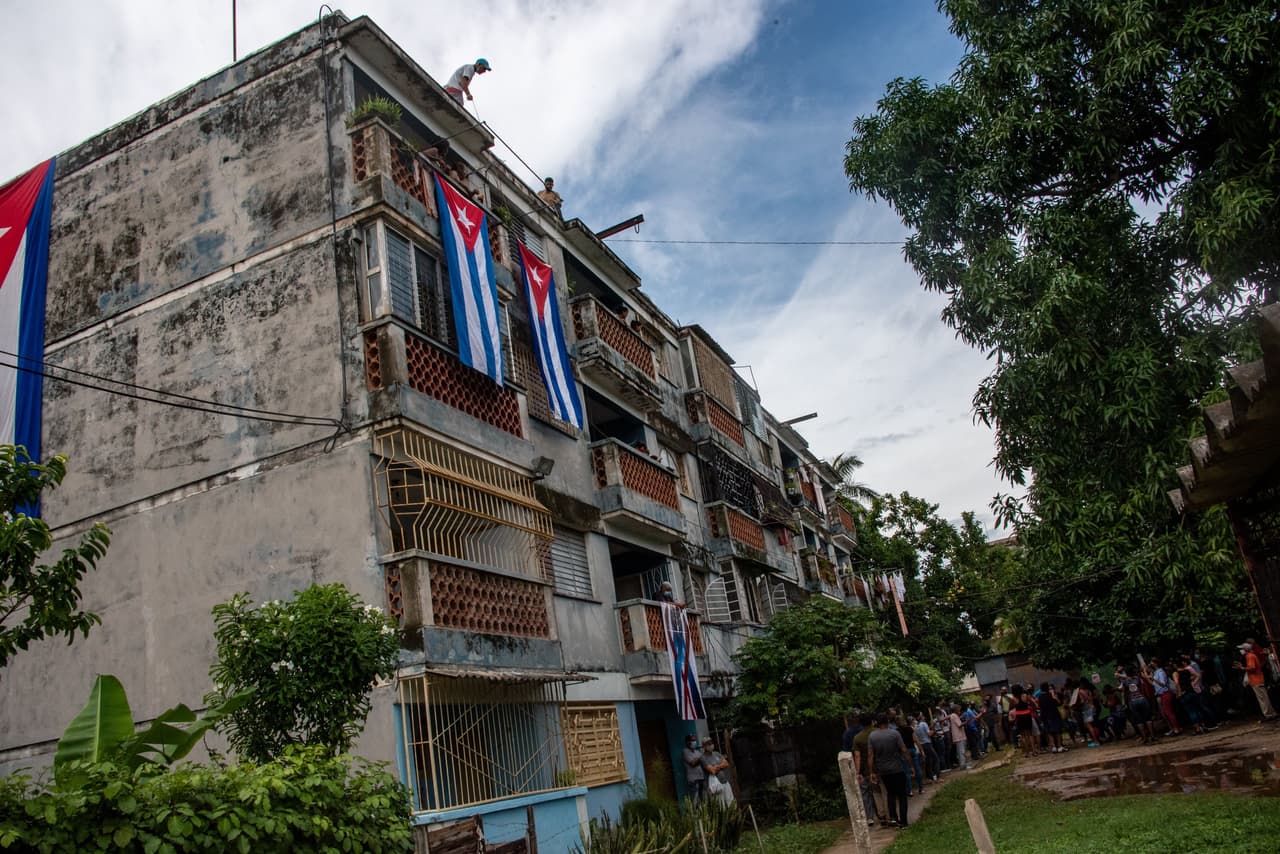 En esta foto se observa a agentes del gobierno, cubriendo con banderas las ventanas de Yunior García Aguilera. Abajo, miembros del partido comunista realizan un acto de repudio en contra del líder del grupo Archipiélago.