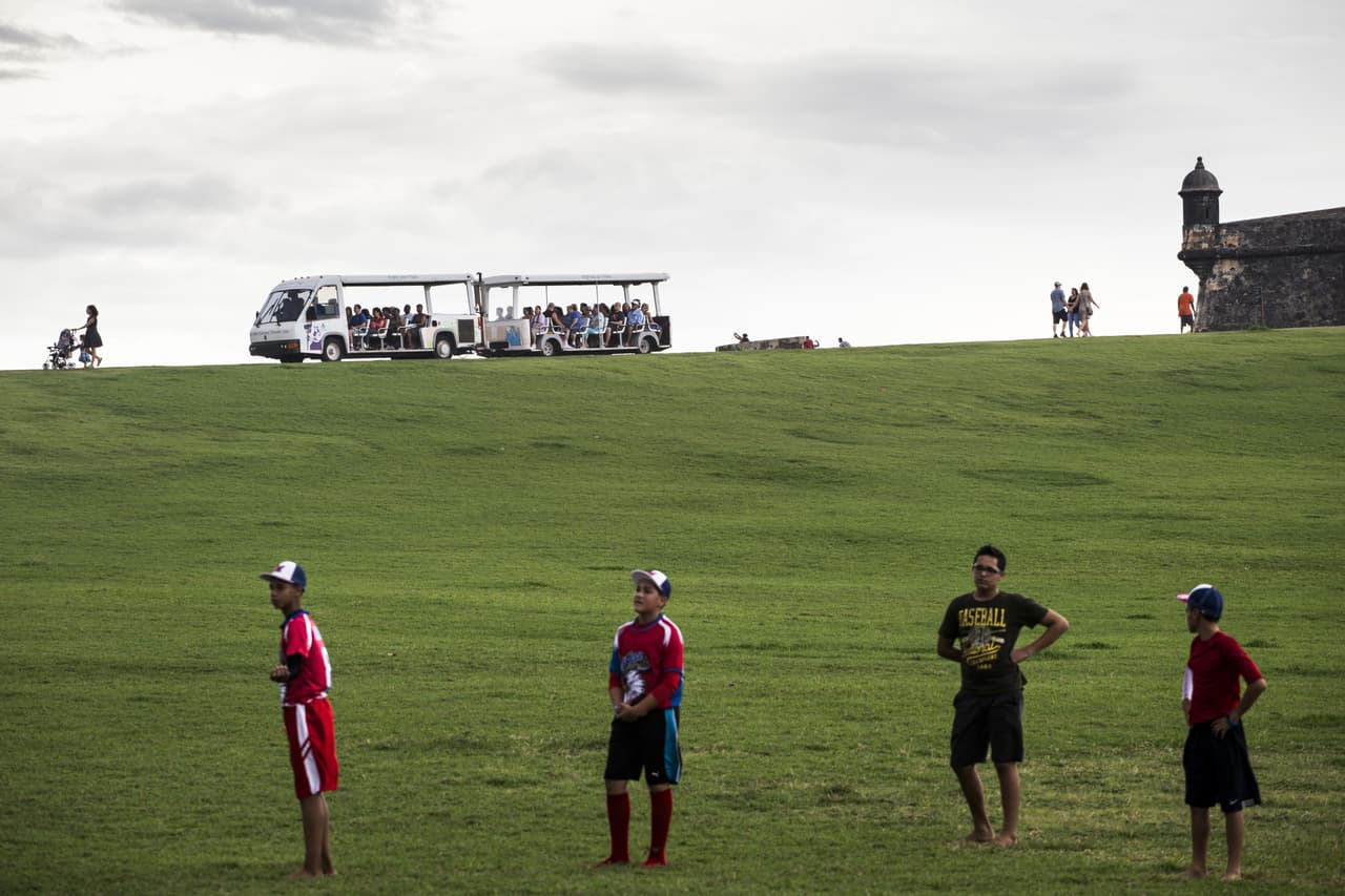 Niños toman una pausa durante un juego de béisbol mientras un transporte lleva turistas al castillo San Felipe El Morro en San Juan. 1 de mayo de 2016.