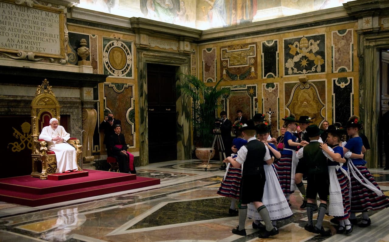 Niños vestidos con trajes tradicionales bávaros bailan frente al Papa Benedicto XVI durante las celebraciones de su cumpleaños número 85 del Pontífice en el salón Clementino de El Vaticano. El papa pasó su cumpleaños entonces con su hermano, Georg, quien falleció en 2020 y quien no quedó exento de escándalos 
<a href="https://www.univision.com/noticias/pederastia/mas-de-500-ninos-fueron-victimas-de-abusos-en-un-coro-catolico-en-alemania-en-mas-de-45-anos-segun-informe" target="_blank">cuando en 2017 estallaron las acusaciones</a> de abuso sexual dentro del coro que dirigía.