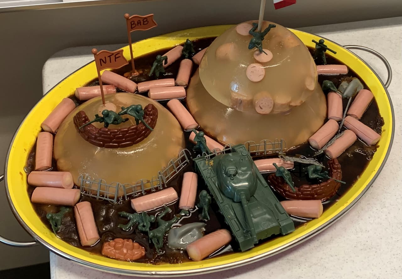 Un diorama de batalla muy poco convencional servido en una bandeja amarilla. La escena incluye un "campo de batalla" de gelatina y salsa de soja, perritos calientes rebanados, un tanque de juguete y soldados de plástico verde, con dos pequeñas banderas que dicen "NTF" y "BAB".